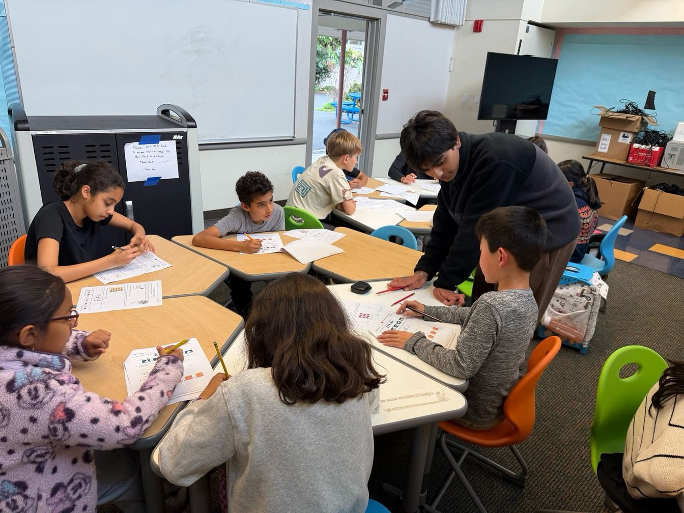 Devansh leaning over a table of younger students working through Math Kangaroo practice problems, with worksheets and pencils spread across the desks.